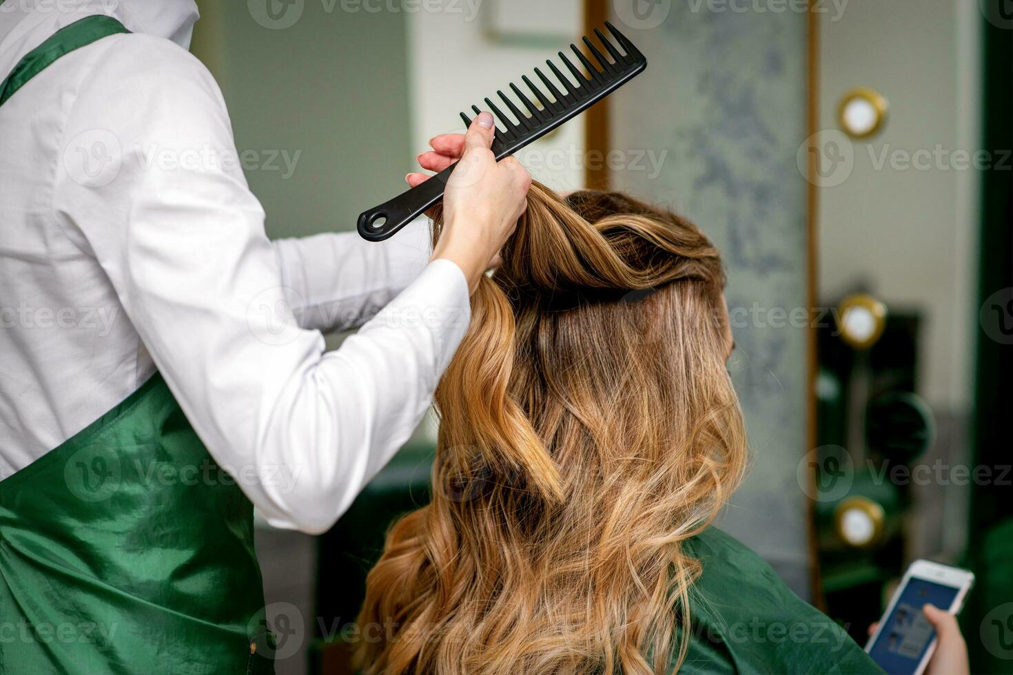 un' femmina parrucchiere è pettinatura il lungo Marrone capelli di un' giovane donna a un' salotto. foto