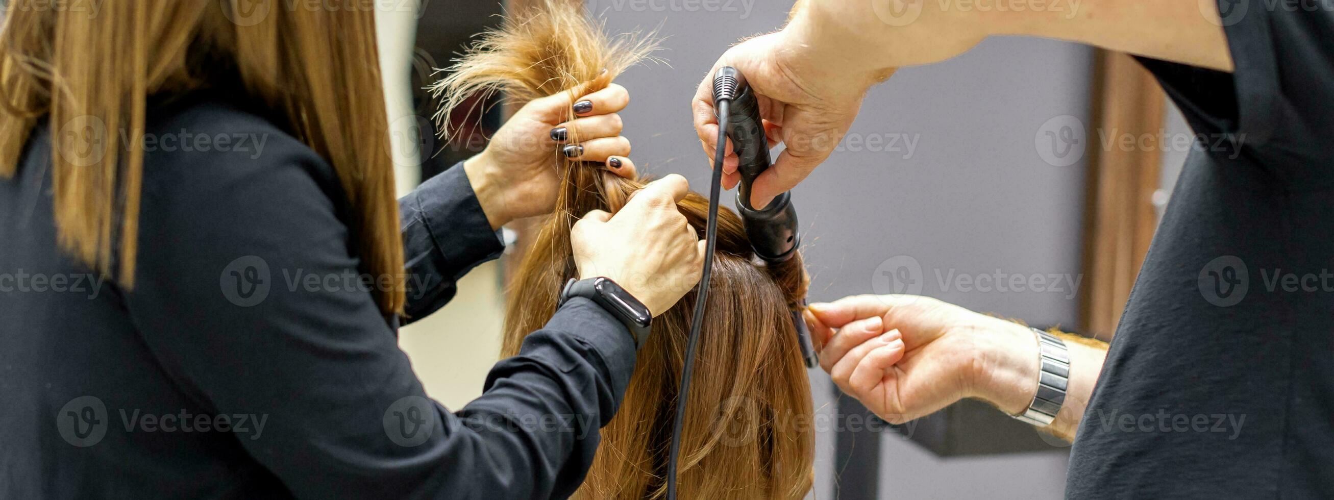 il posteriore Visualizza di Due parrucchieri siamo arricciatura capelli per un' giovane donna con elettrico capelli ferro nel un' bellezza salone. foto