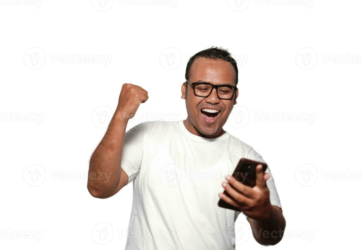 eccitato asiatico uomo con bicchieri indossare bianca maglietta sorridente mentre Tenere il suo Telefono, isolato di bianca sfondo foto