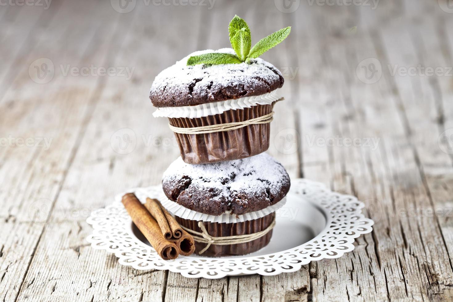 Muffin al cioccolato fondente con zucchero in polvere, bastoncini di cannella e foglia di menta su piastra bianca su tavola in legno rustico. foto