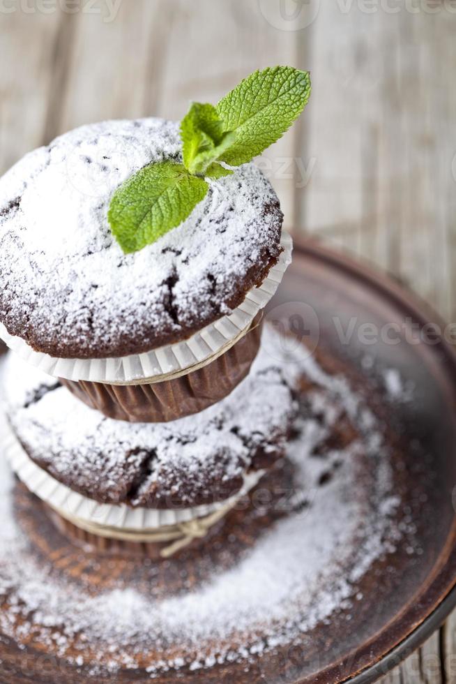 muffin al cioccolato fondente freschi con zucchero in polvere e foglia di menta su piastra marrone su tavola in legno rustico. foto