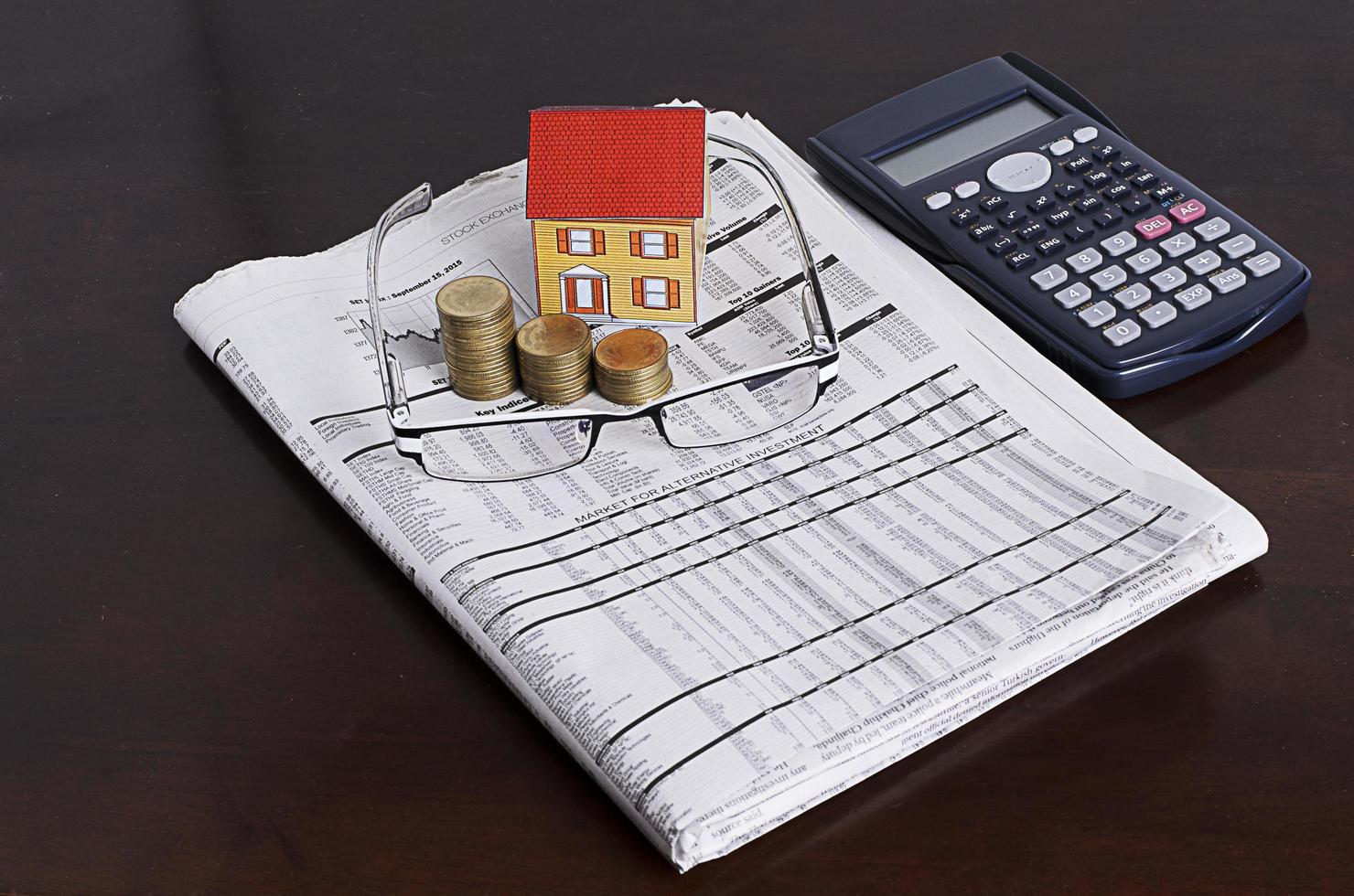 concetto di prestiti ipotecari con casa di carta con pila di monete, occhiali e giornali, calcolatrice foto