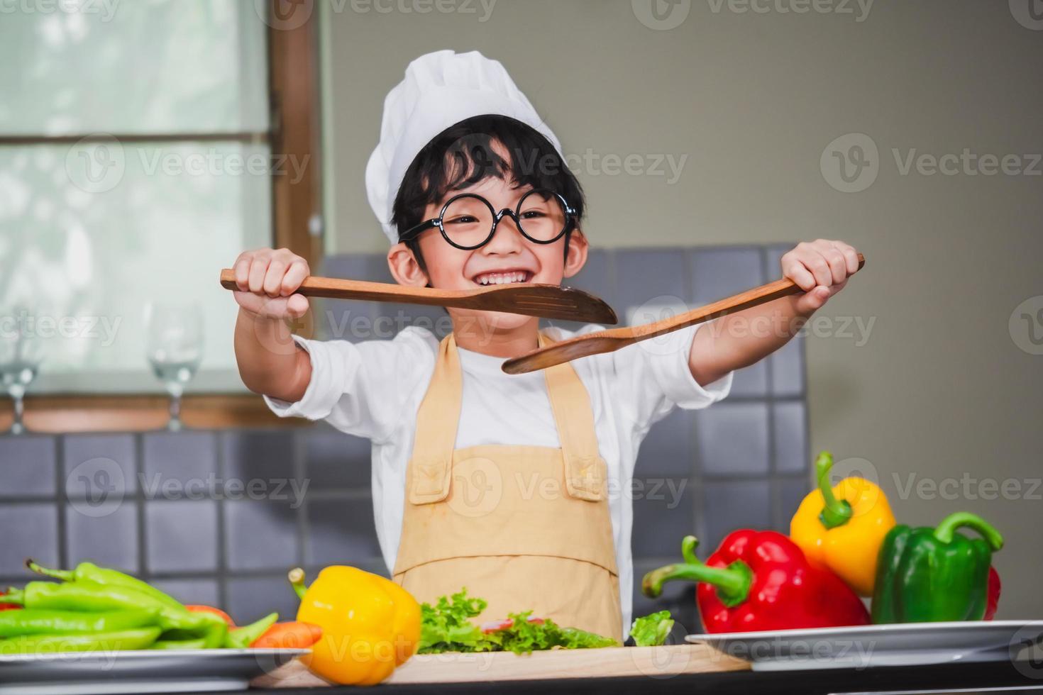 ragazzo asiatico figlio che cucina insalata cibo holdind cucchiaio di legno con verdura che tiene pomodori e carote, peperoni sul piatto per famiglia felice cucinare cibo godimento stile di vita cucina in casa foto