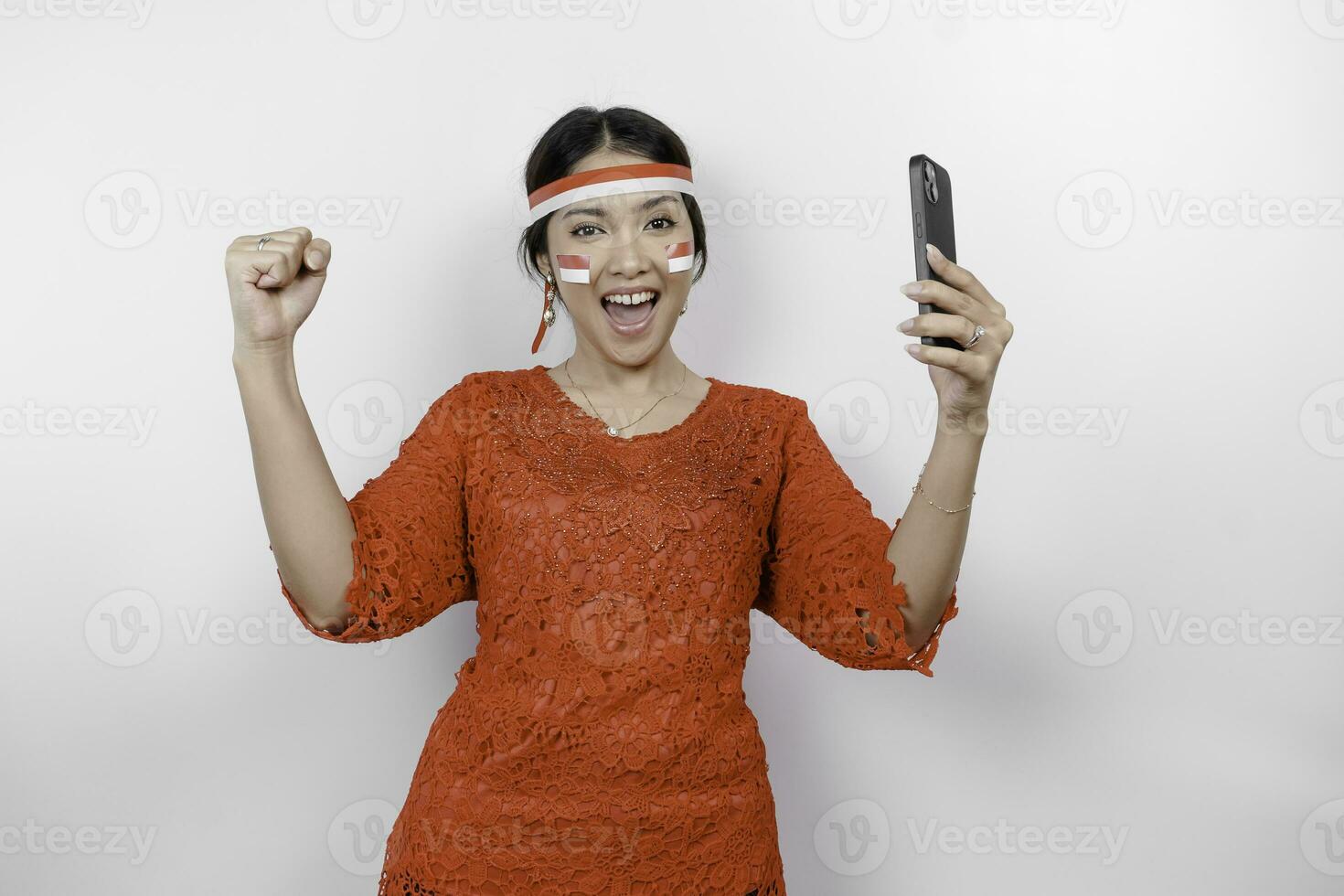 un' giovane asiatico donna con un' contento riuscito espressione mentre Tenere sua Telefono e indossare rosso kebaya, bandiera fascia per capelli isolato di bianca sfondo. dell'Indonesia indipendenza giorno concetto. foto