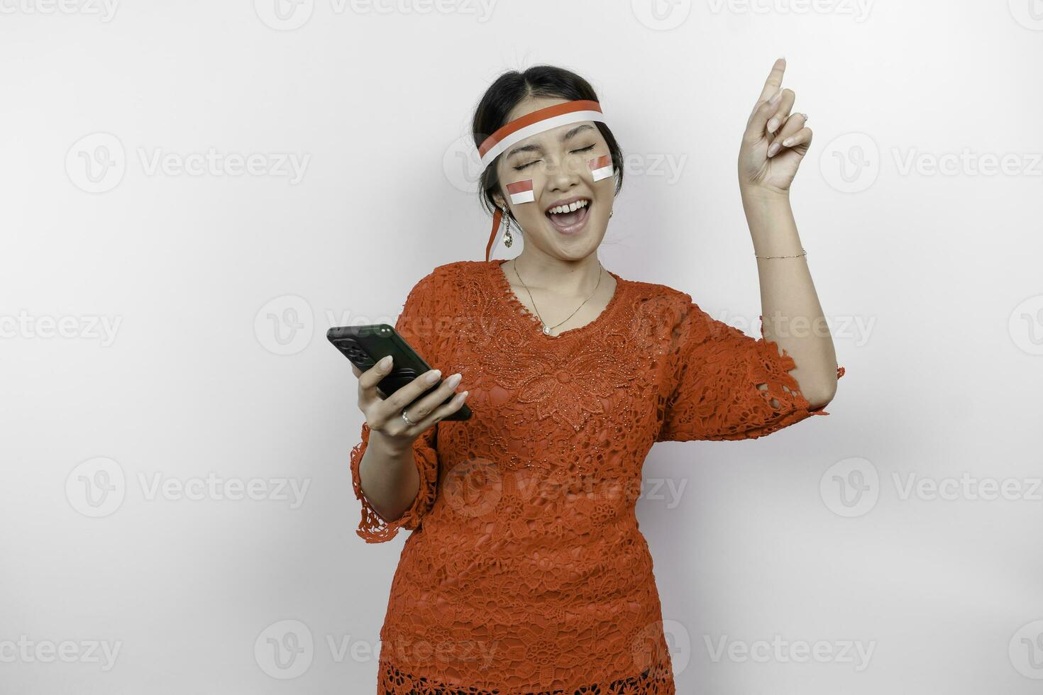 un' contento asiatico donna indossare rosso kebaya e fascia per capelli, Tenere sua Telefono, e puntamento copia spazio su superiore di suo, isolato di bianca sfondo. dell'Indonesia indipendenza giorno foto