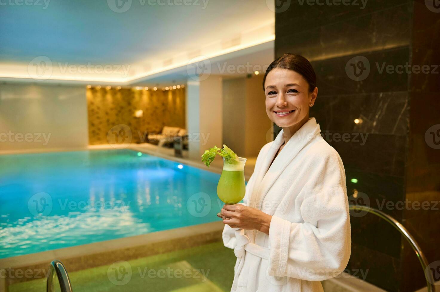 affascinante donna nel bianca accappatoio, detiene un' bicchiere con delizioso salutare appena spremuto vitamina succo nel sua mani, sorrisi guardare a telecamera, in piedi di il vasca idromassaggio a benessere terme centro sala la zona foto