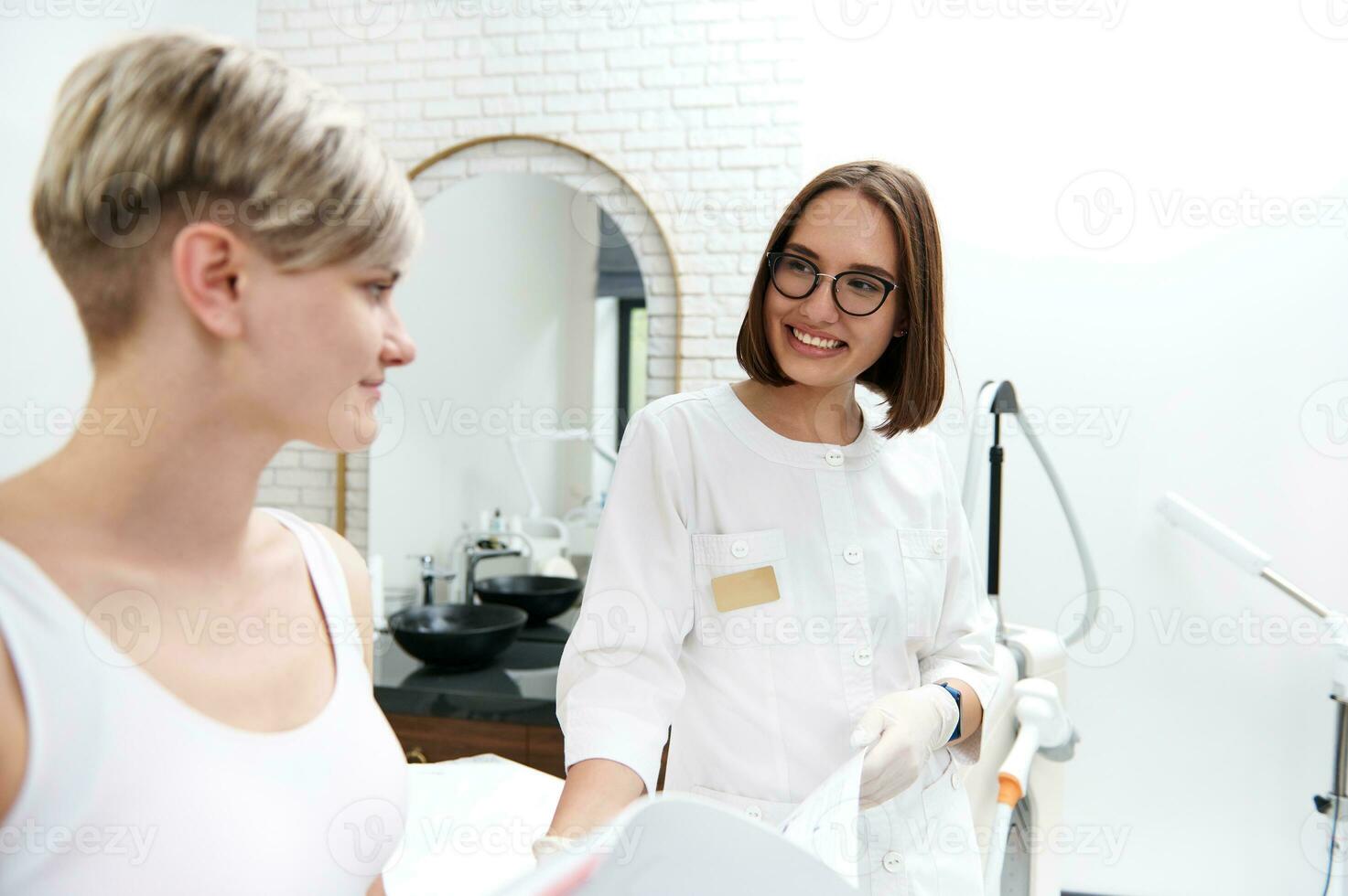sorridente cosmetologo mostrando un' Catalogare di prezzi per cosmetico procedure per un' giovane cliente g durante un' consultazione a un' bellezza terme salone foto