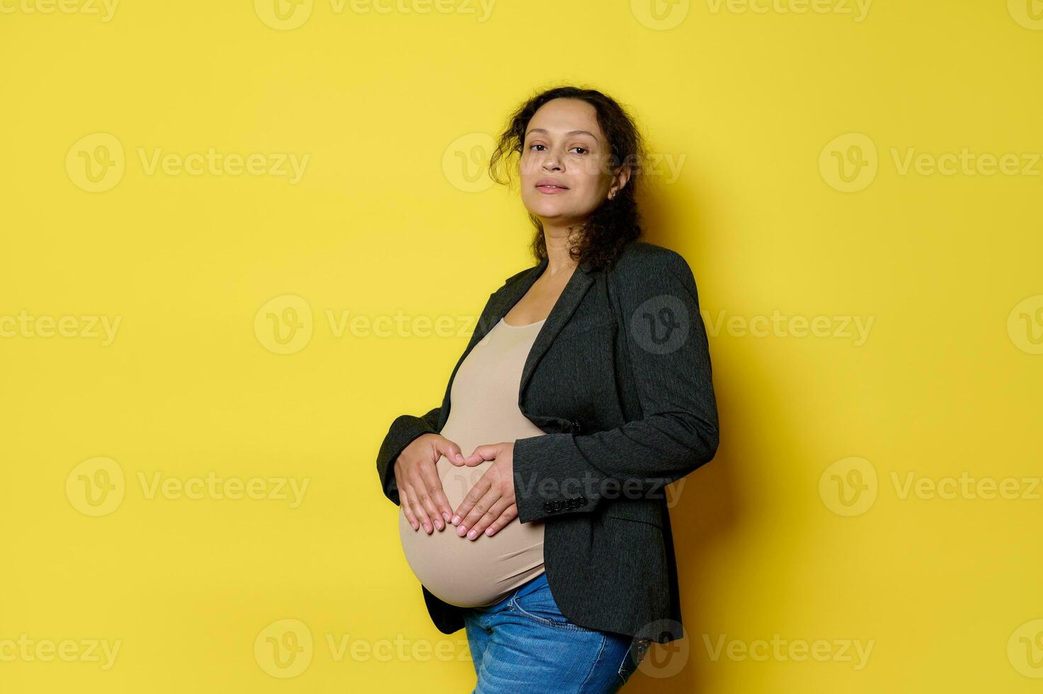 bellissima incinta donna aspettandosi bambino, formatura un' cuore forma con sua dita su sua gonfiarsi, isolato giallo sfondo foto