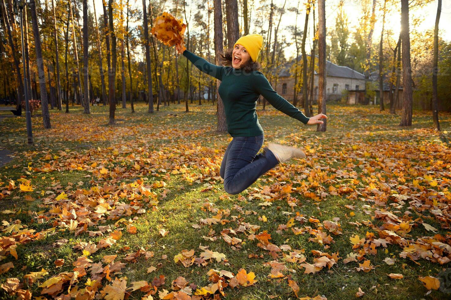 bellissimo donna nel caldo verde Maglione, giallo di lana e casuale denim cappello salto alto con un' mazzo di bellissimo asciutto autunno acero le foglie nel mani, godendo bellissimo autunno giorno a tramonto nel parco foto