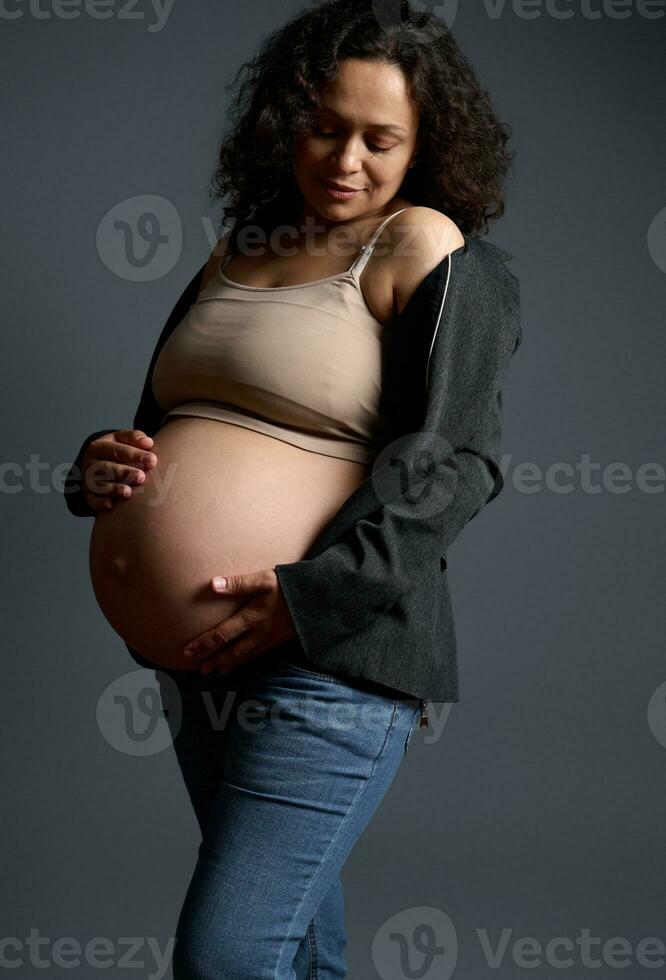 ritratto di bellissima incinta donna aspettandosi bambino, mettendo mani su sua grande pancia nel il nono mese di contento gravidanza foto