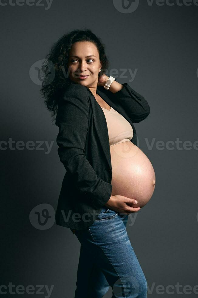 sfocatura arte. autentico latino gravido donna, delicatamente tocchi sua incinta gonfiarsi, sorrisi guardare a parte, moda grigio sfondo foto