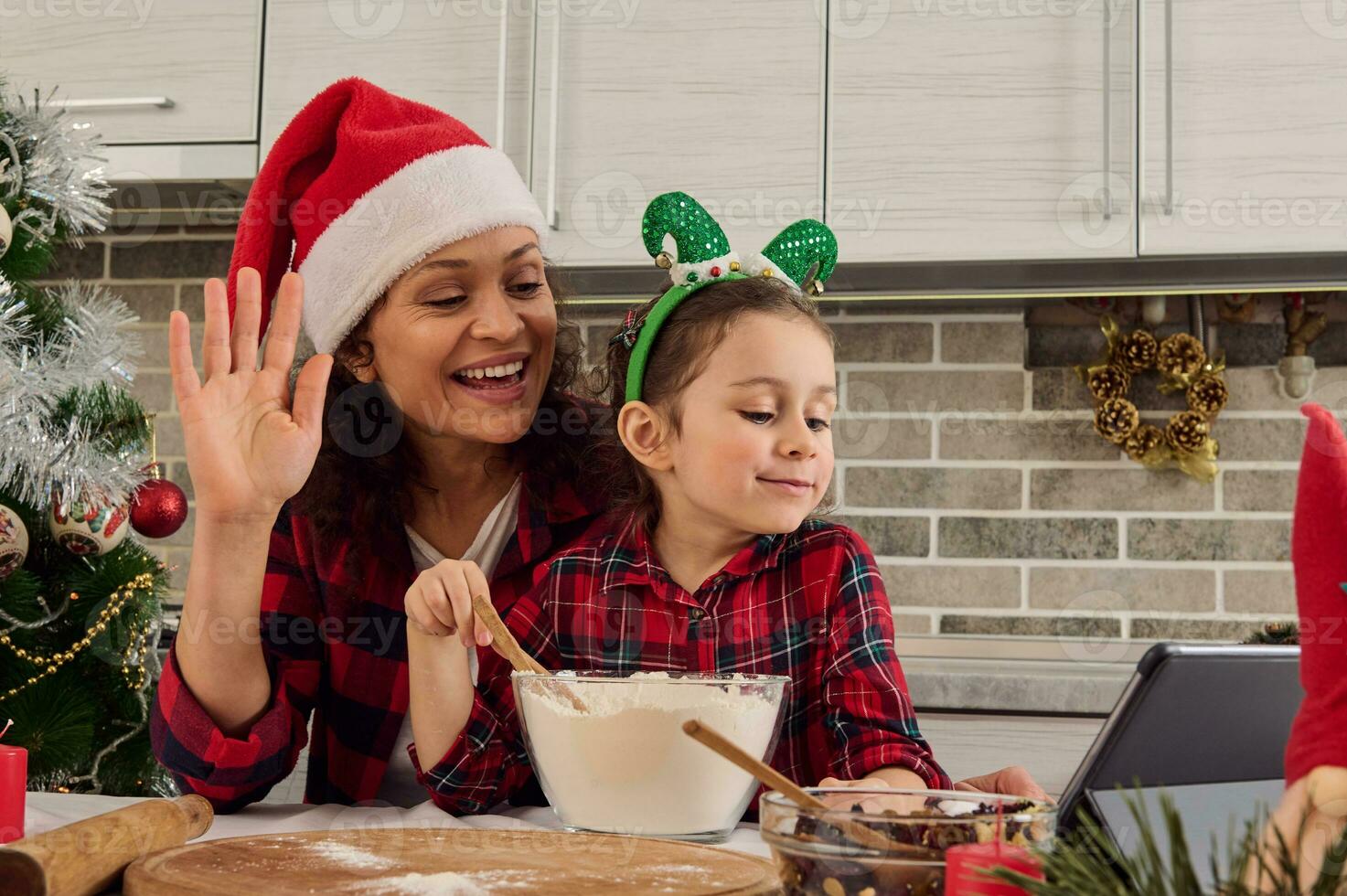 contento mamma nel Santa cappello e sua bellissimo poco figlia saluto mentre parlando di video chiamata , registrazione video per loro cibo culinario video blog mentre cucinando insieme Natale Pasticcino a casa cucina foto