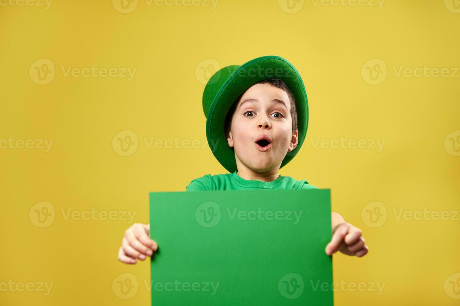 sorpreso ragazzo nel verde irlandesi fata cappello pose spirito un' verde foglio di carta nel il suo mani. santo Patrick giorno concetto. giallo sfondo. copia spazio. foto