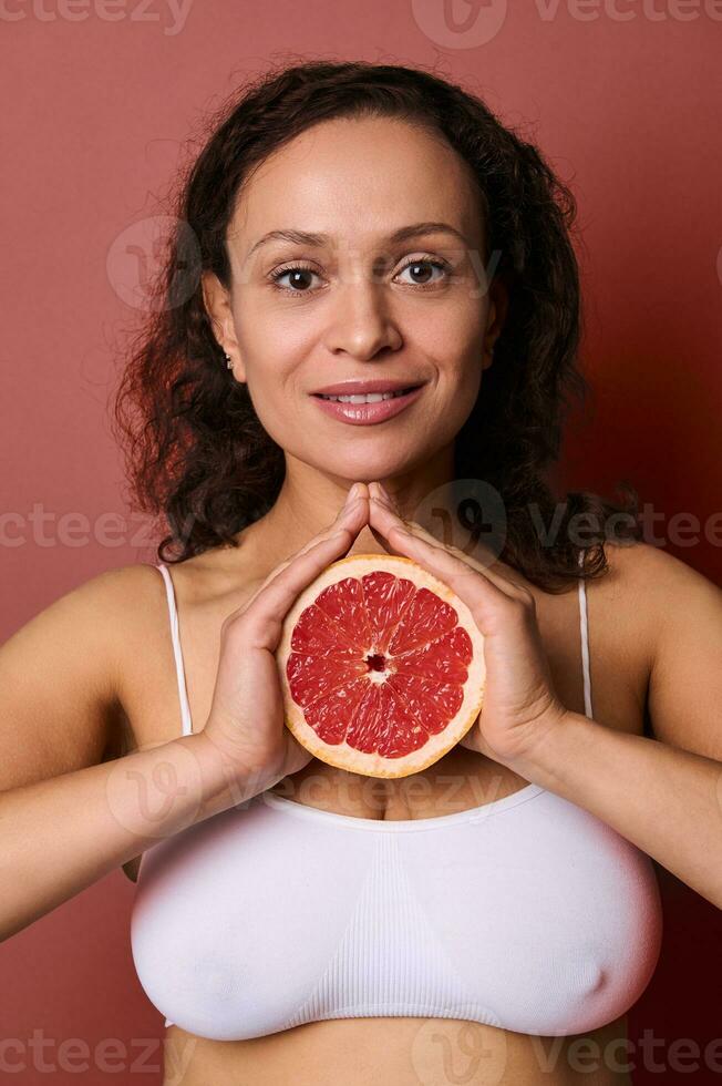 bellezza ritratto di un attraente sorridente mezzo anziano bellissimo donna con Riccio buio capelli capelli, indossare bianca biancheria intima, isolato su luminosa corallo sfondo, Tenere metà di fresco succoso rosso pompelmo foto