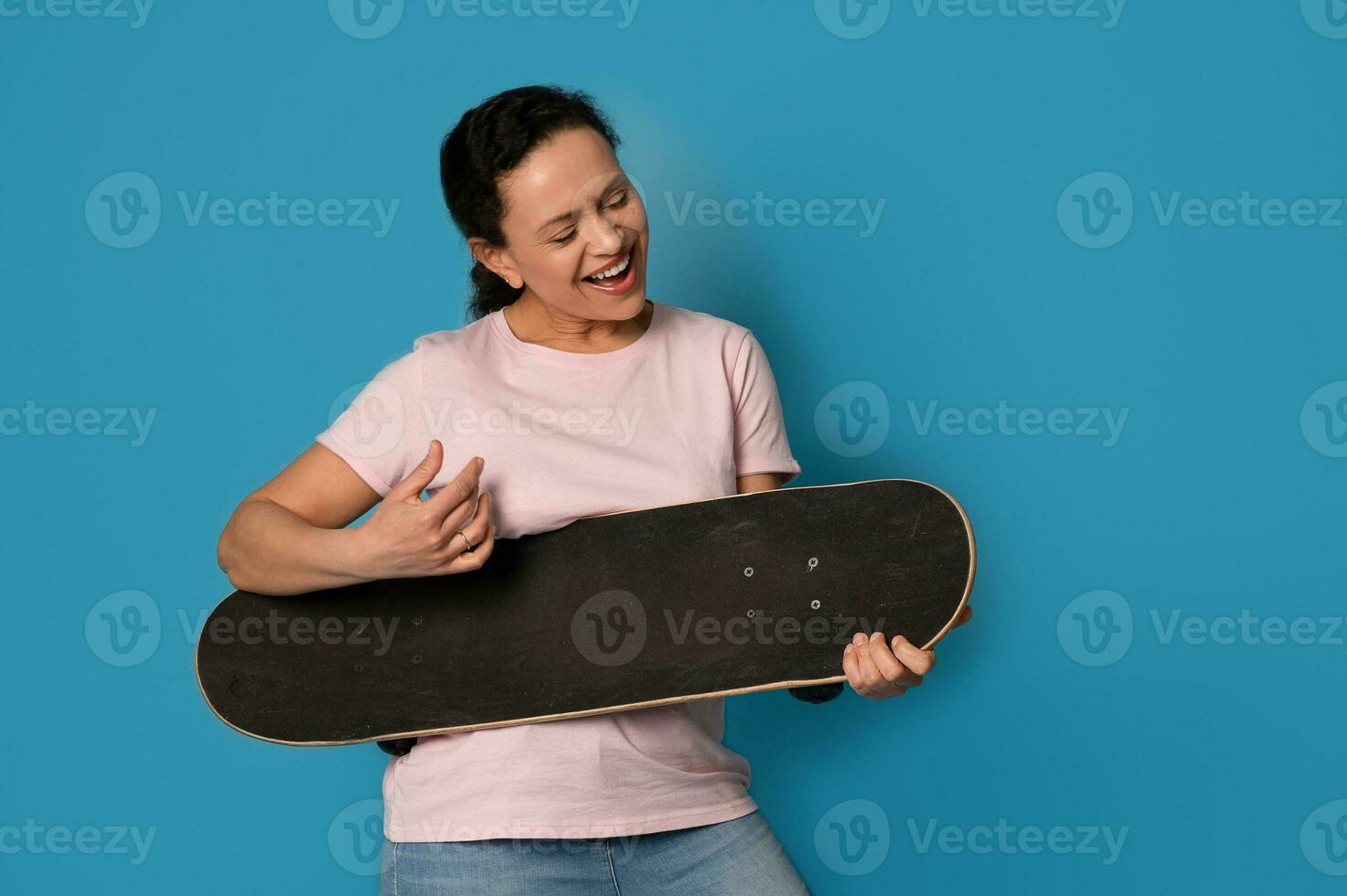 gioioso donna skateboarder Tenere un' skateboard e imitando giocando chitarra, isolato al di sopra di blu sfondo foto