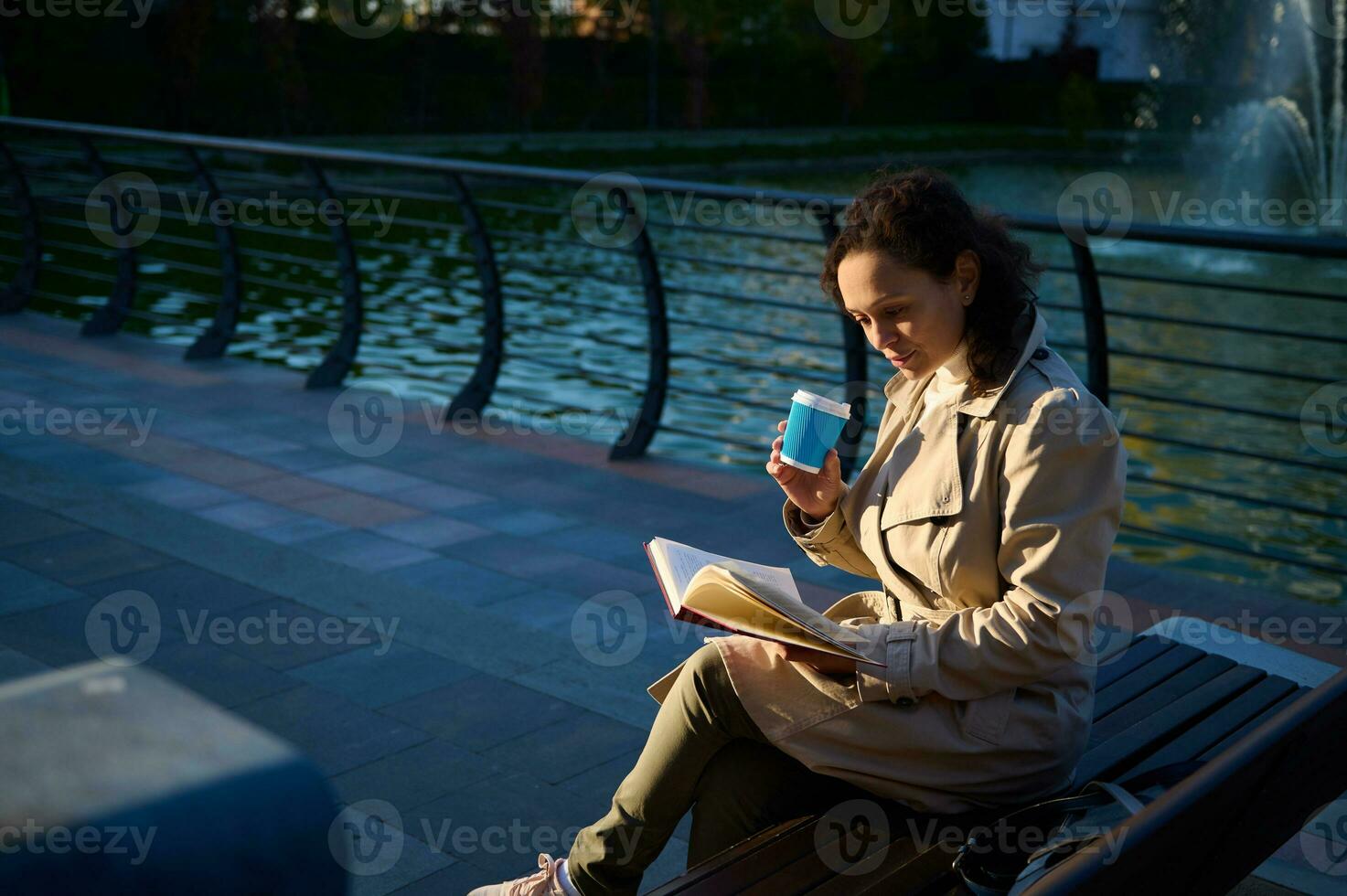 giovane donna nel beige trincea cappotto seduta su un' parco panchina su il lago sfondo , potabile caffè o caldo bevanda nel riciclabile porta via carta tazza e lettura prenotare, godendo riposo a partire dal digitale gadget foto