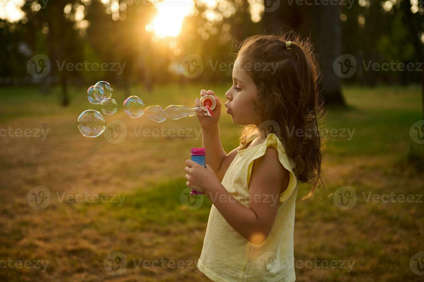 adorabile bambino bambino ragazza, soffiaggio sapone bolle a tramonto, godendo piacevole tempo all'aperto nel il prato. di sole raggi autunno attraverso trasparente bolla sfere con iridescente riflessi. foto