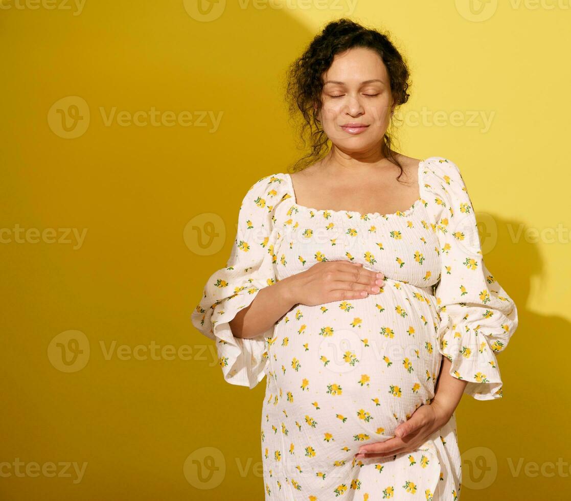 autentico incinta donna Tenere sua gonfiarsi, vestito nel estate vestire, in posa con occhi chiuso su giallo studio sfondo foto