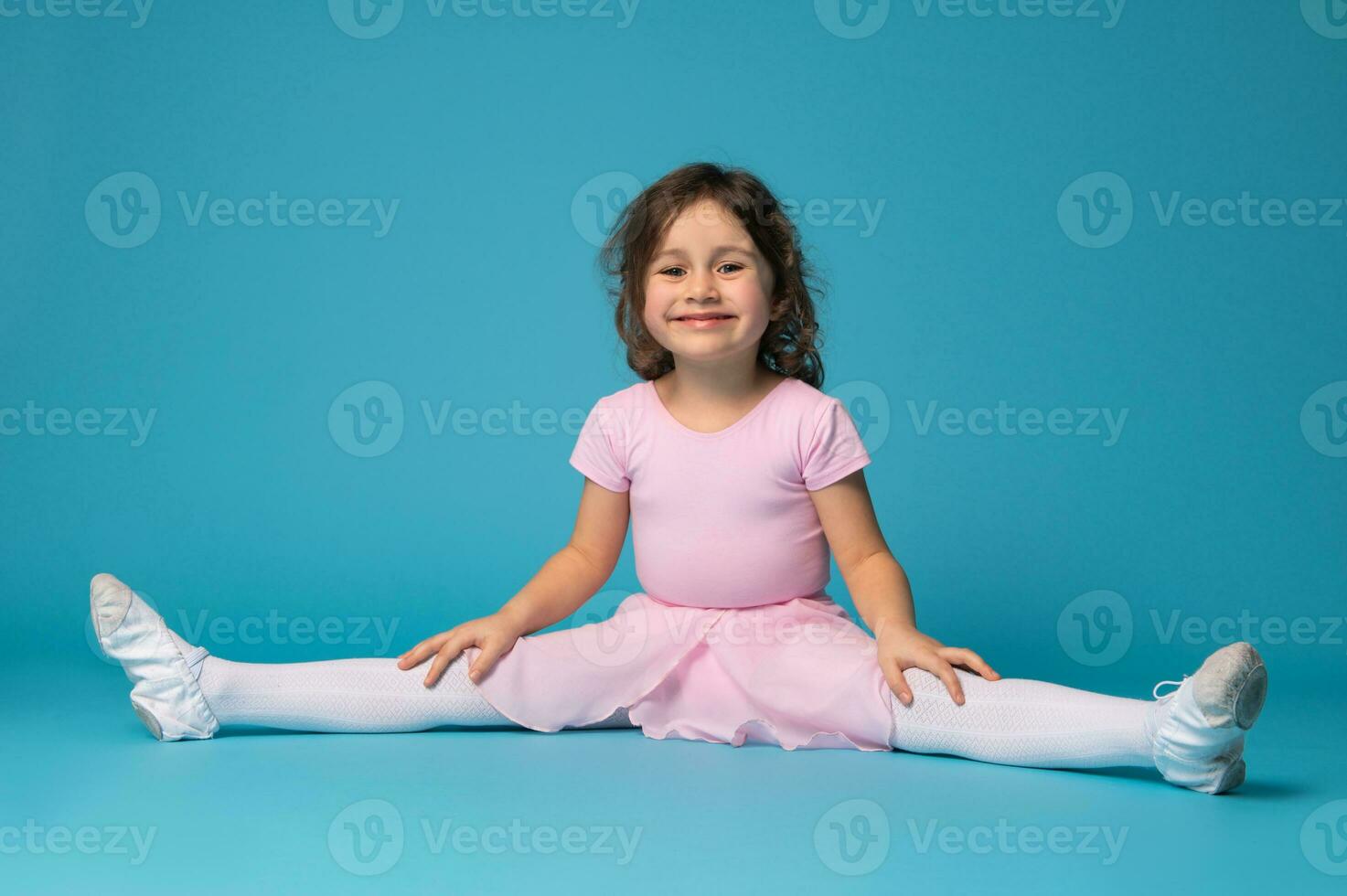 bellissimo poco ragazza ballerina pratiche allungamento sua gambe e carino sorrisi per il telecamera, isolato su blu sfondo foto