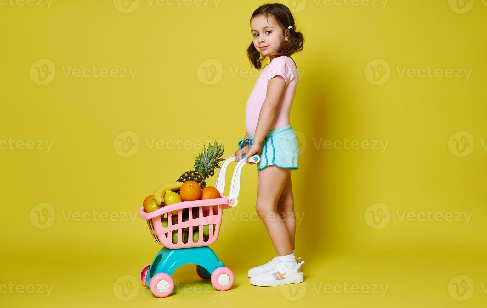 pieno lunghezza ritratto di carino ragazza nel rosa body e blu estate pantaloncini con shopping carrello pieno di frutta in posa contro giallo sfondo foto