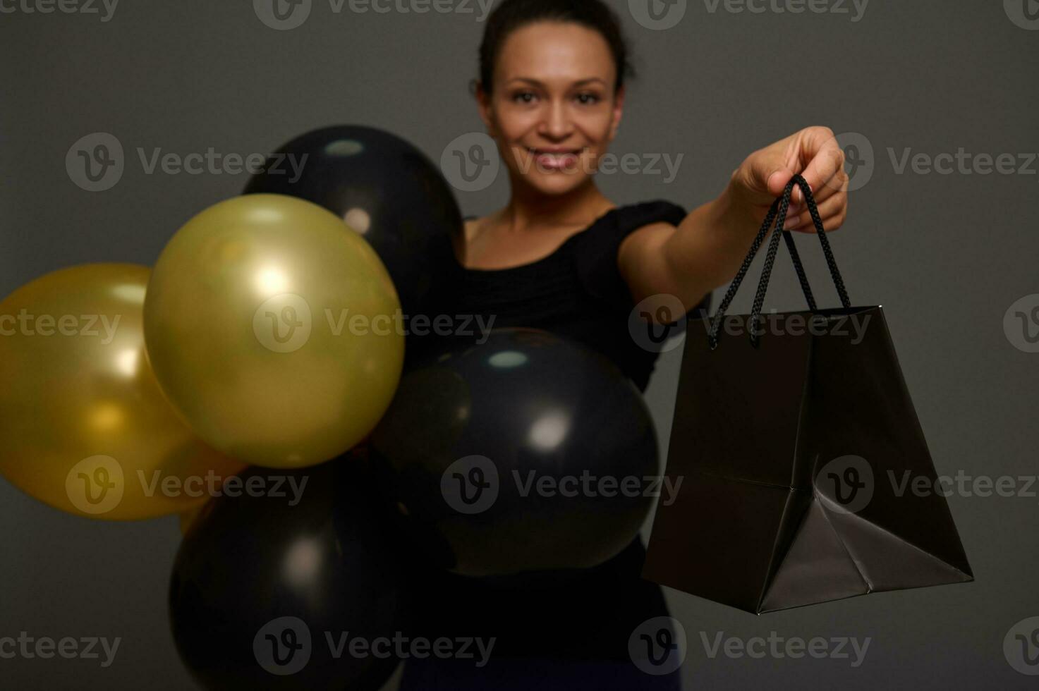 messa a fuoco su il mano Tenere un' nero shopping Borsa. sfocato sorridente donna con oro nero aria palloncini e pacchetto su grigio sfondo con copia spazio per nero Venerdì i saldi annuncio pubblicitario foto