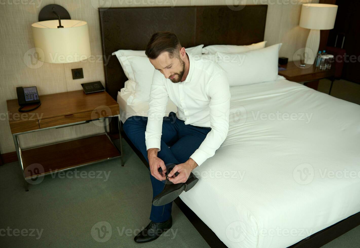 alto angolo Visualizza di un' bello europeo uomo d'affari nel attività commerciale abbigliamento legatura il suo lacci delle scarpe, seduta su bordo di letto nel Hotel Camera da letto, rilassante dopo incontri durante attività commerciale viaggio foto