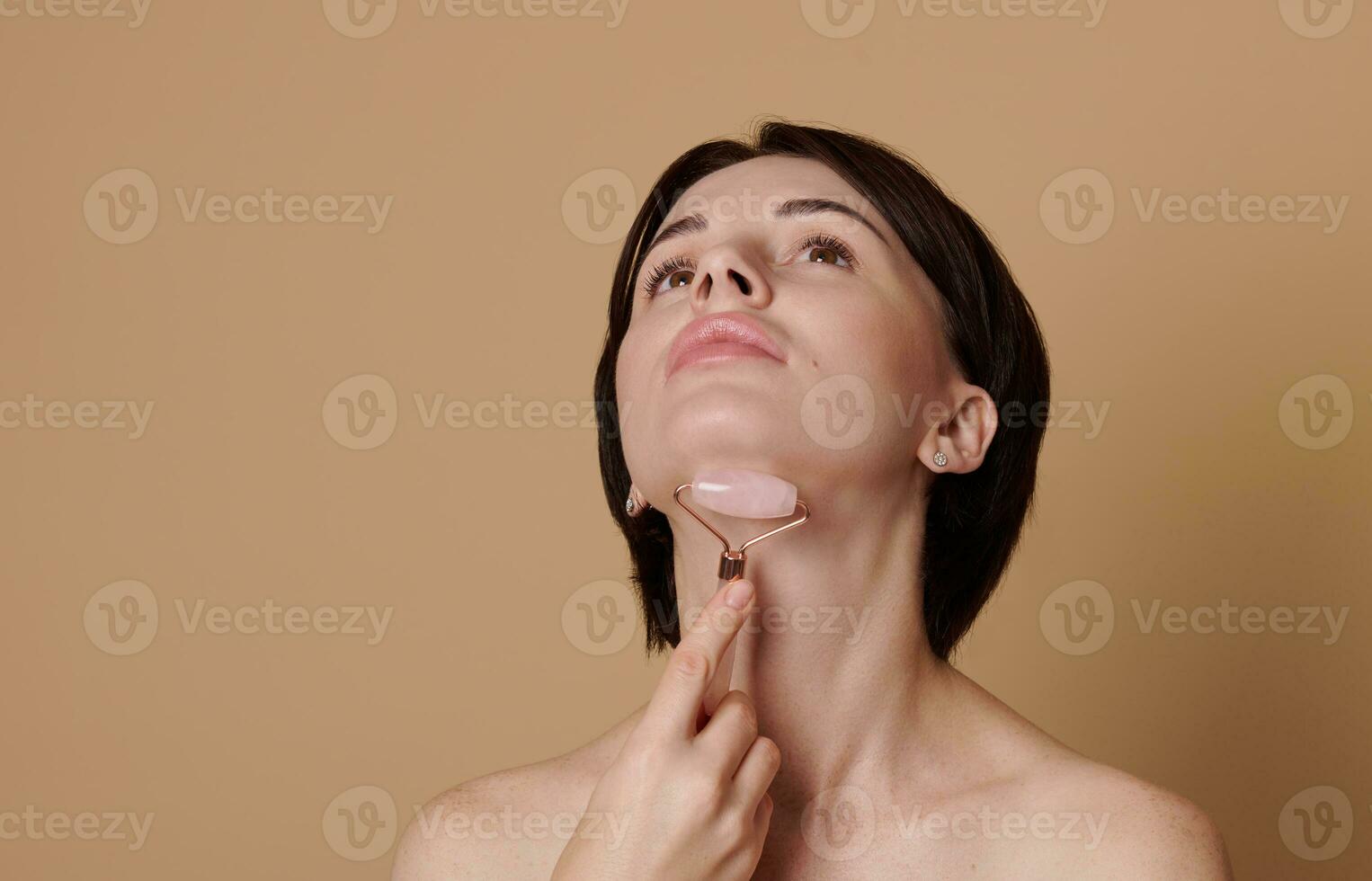 giovane brunetta donna fare un' sollevamento massaggio su sua collo, utilizzando un' giada rullo massaggiatore. bellezza ritratto su beige sfondo con spazio per anno Domini. foto