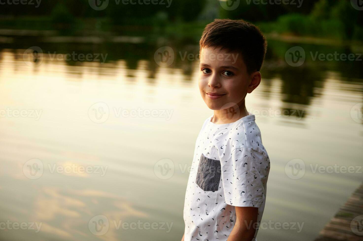 ritratto di un' affascinante ragazzo carino sorridente, guardare a il telecamera su il sfondo di il lago. tramonto e un' linea di alberi siamo riflessa nel il lago acqua foto