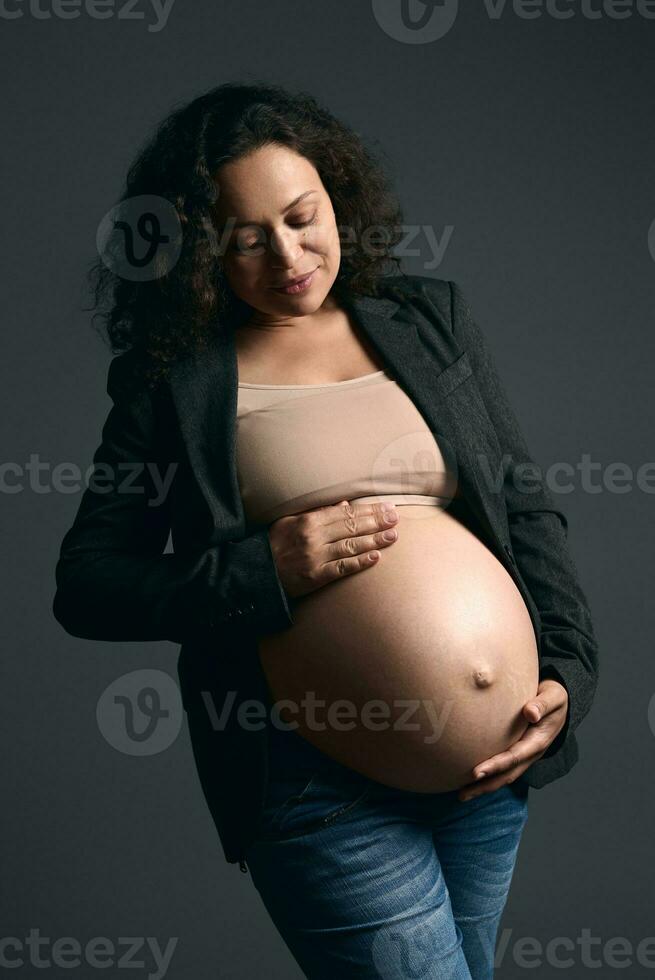 autentico incinta donna nel elegante grigio blazer e blu jeans, sorridente, accarezzando sua spoglio gonfiarsi, su studio sfondo foto