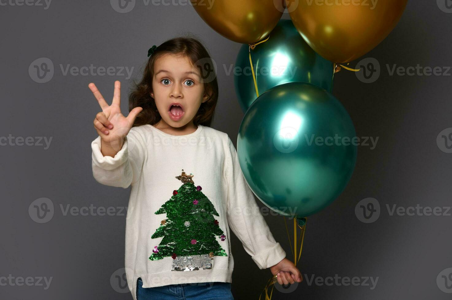 bellissimo ragazza indossare bianca maglione con Natale albero, detiene bellissimo d'oro e verde metallico aria palloncini nel sua mano, Spettacoli dita guardare a telecamera, pose contro grigio sfondo, copia spazio foto