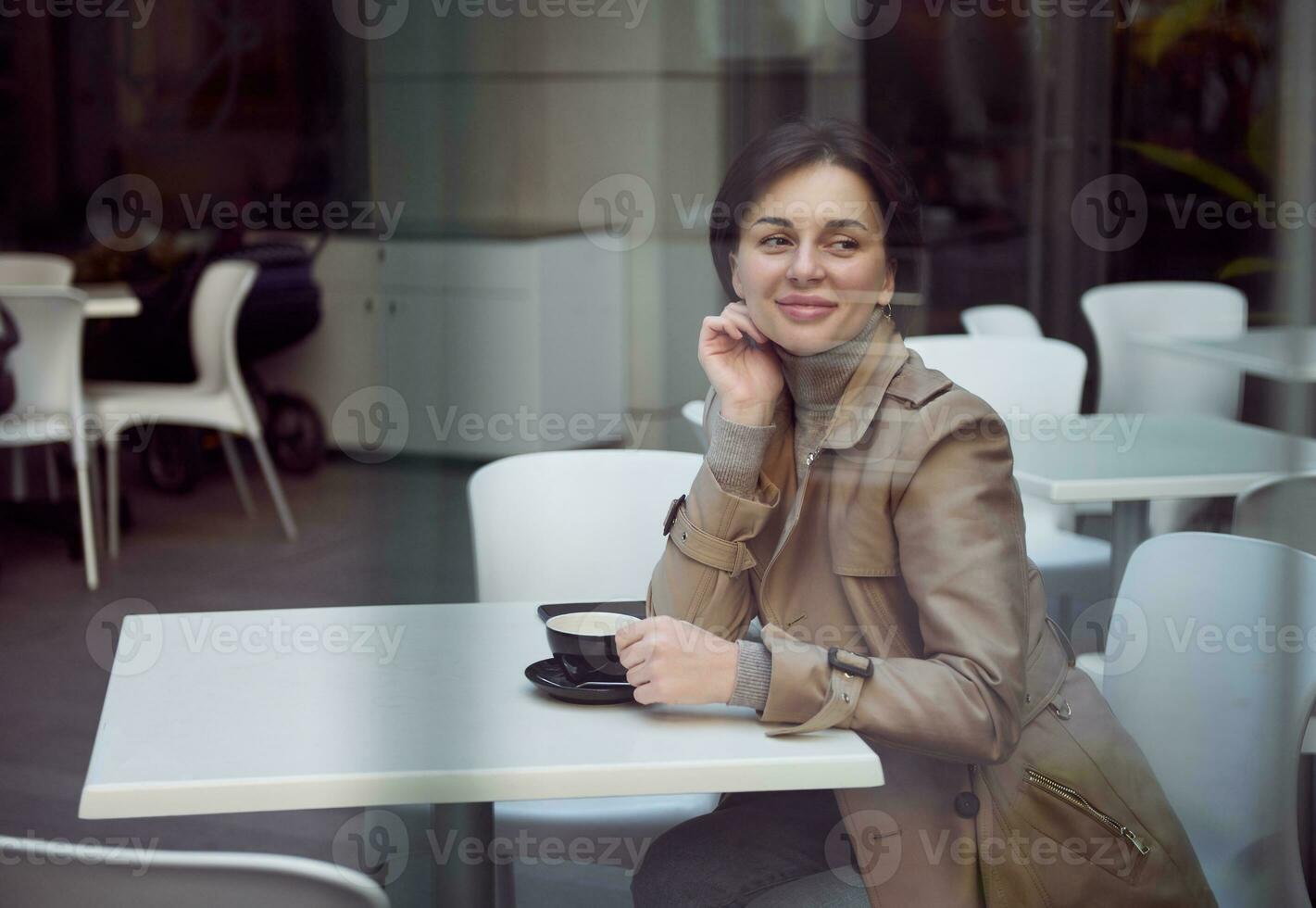 bellissimo attraente sbalorditivo brunetta donna con un' elegante corto taglio di capelli nel un' beige trincea cappotto seduta a un' tavolo all'aperto e gode mattina caffè con un' sognante Guarda foto