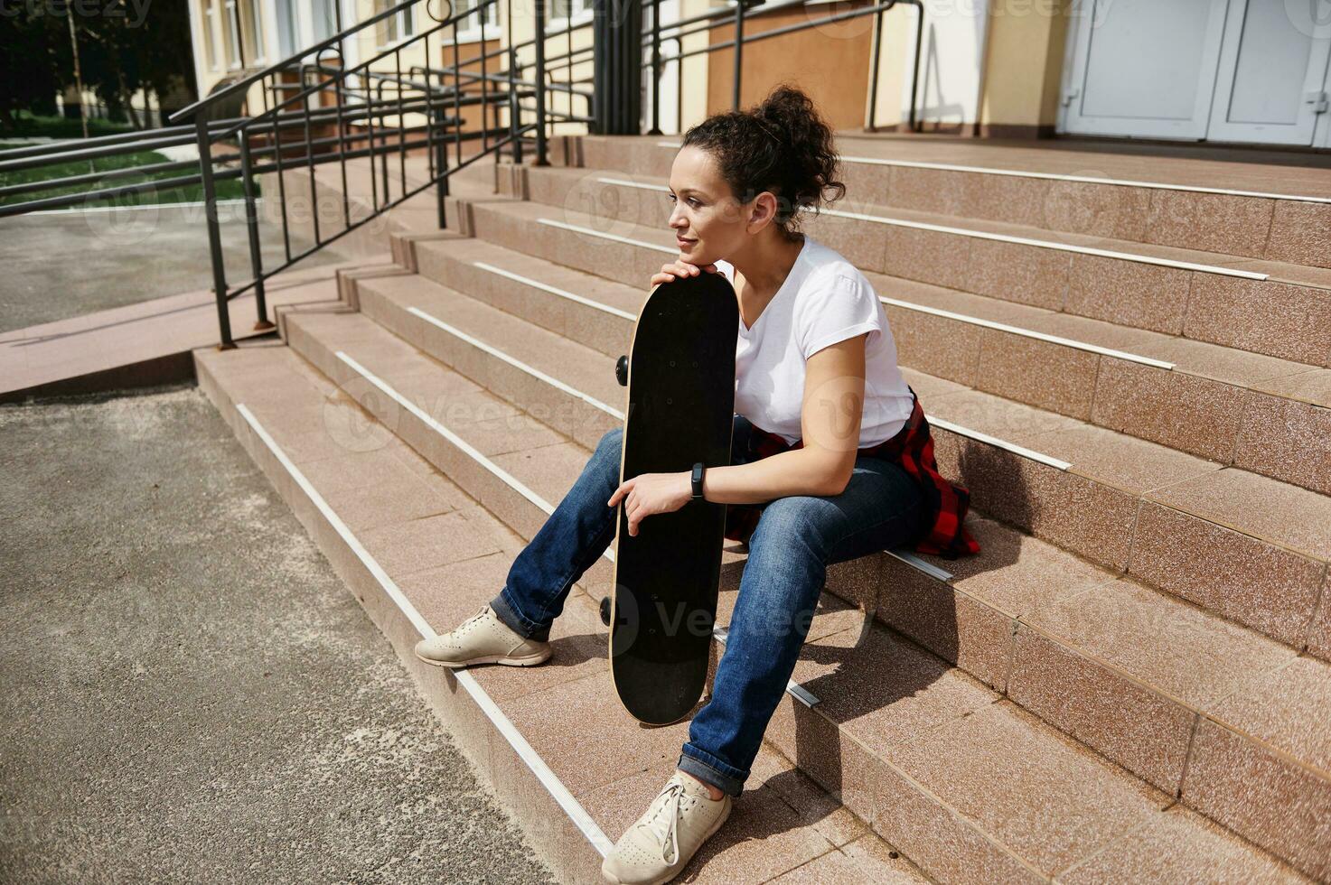 bellissimo giovane africano americano donna con skateboard seduta su passaggi di irriconoscibile edificio foto