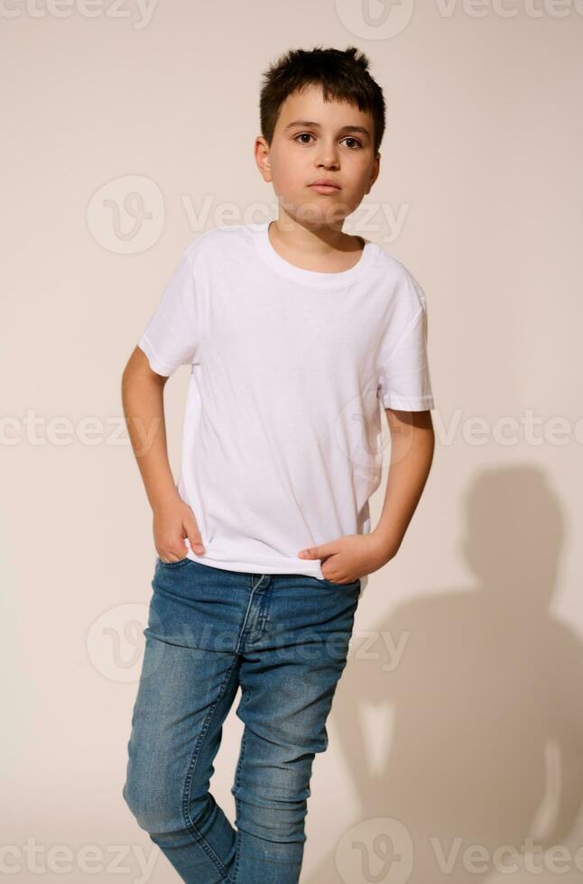 autentico ispanico adolescenziale ragazzo nel bianca maglietta e blu jeans, mettendo il suo mani nel tasche, pensieroso guardare lontano foto