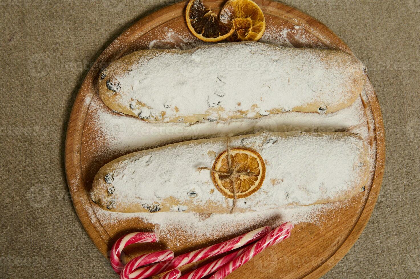 piatto posare cibo composizione di tradizionale Tedesco pane Stollen al forno per Natale, ornato con un' asciutto arancia fetta e un' corda , caramella canne su un' di legno tavola su un' biancheria tovaglia foto