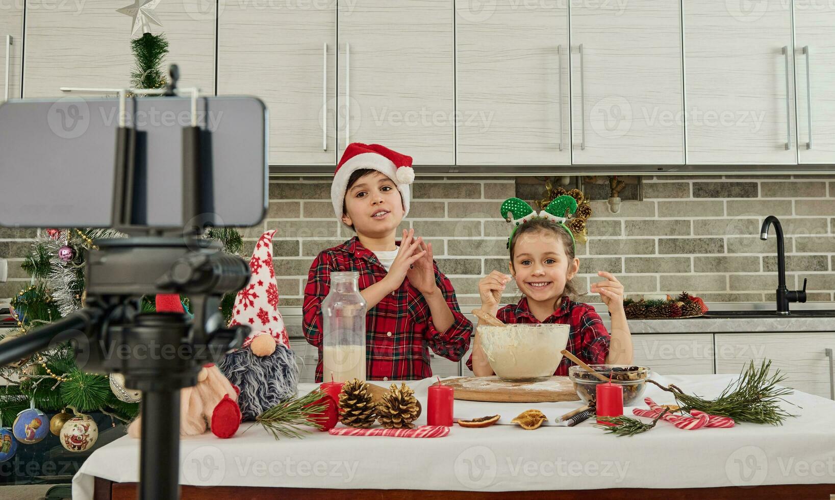 bellissimo bambini chef, carino ragazza e bello ragazzo indossare Santa cappello guardare a telecamera nel smartphone montato su treppiedi, registrazione video per vlog, cucinando Natale pane insieme nel cucina foto