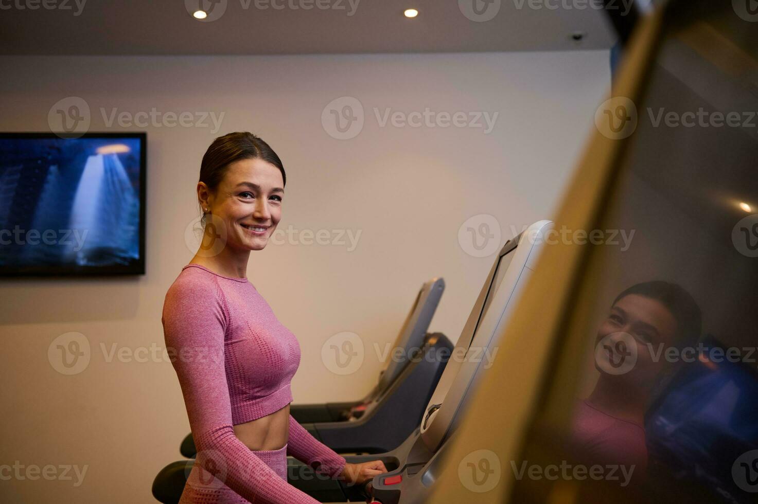 lato Visualizza di un' mezzo anziano e allegro donna nel abbigliamento sportivo in esecuzione su un' routine a Palestra e sorrisi guardare a telecamera. cardio allenamento. foto