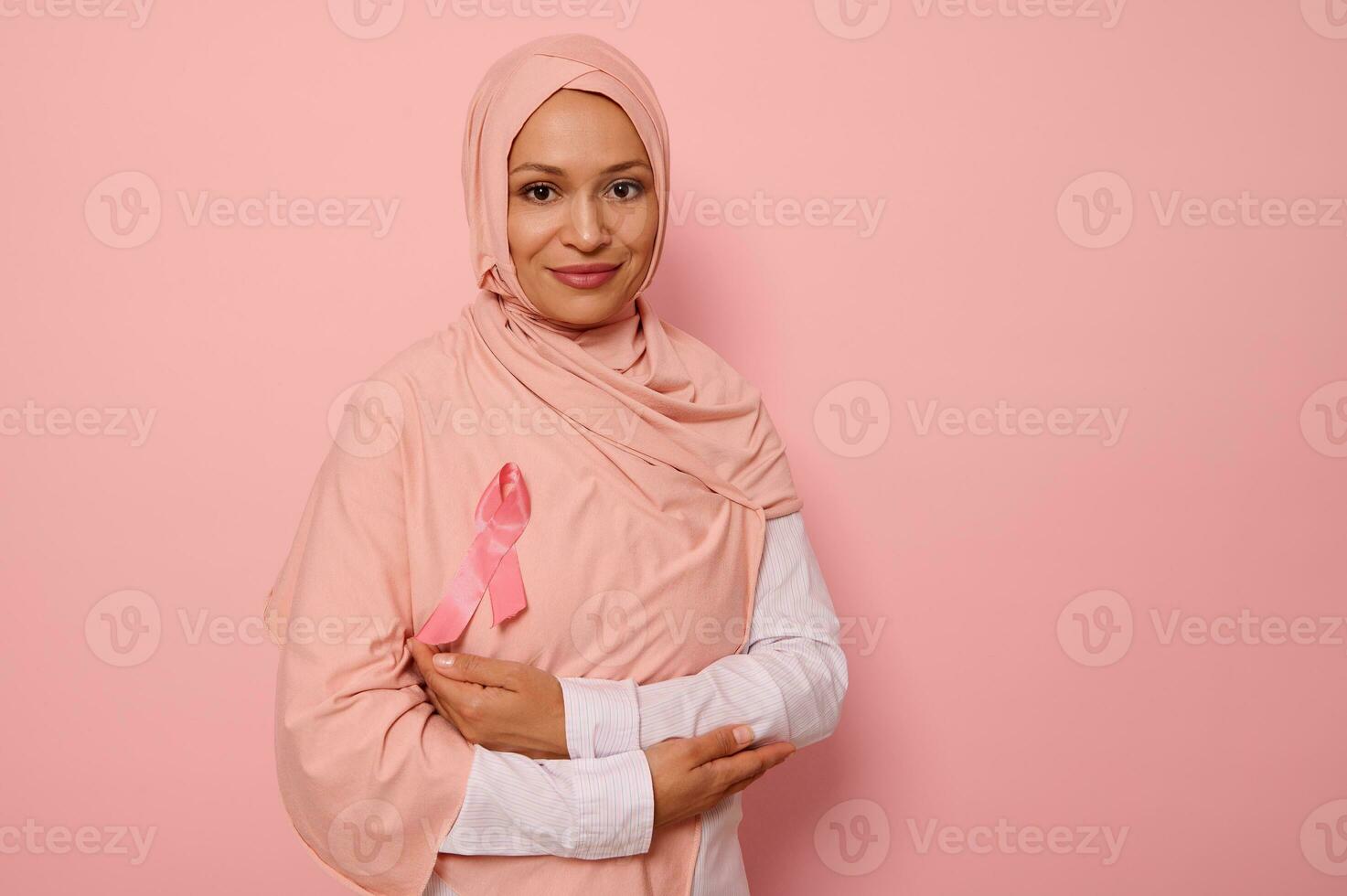 bellissima musulmano donna con coperto testa nel hijab, sta su rosa sfondo con attraversato braccia su il petto e indossare un' rosa nastro, simbolo di Seno cancro consapevolezza giorno. medico concetto, copia spazio foto