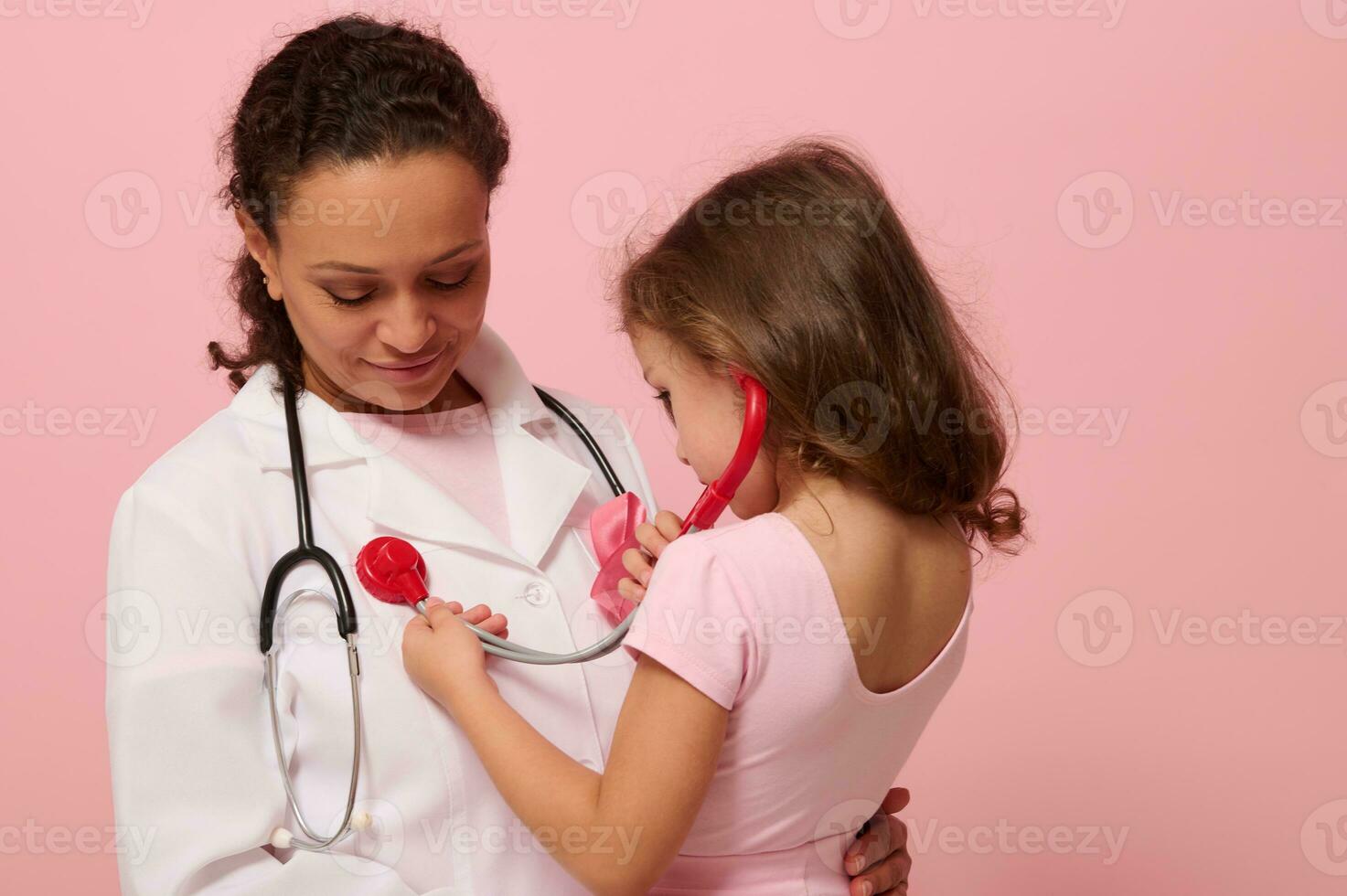 adorabile 4 yo bambino ragazza giocando medico, utilizzando stetoscopio, ascoltando per il petto e polmoni respirazione di medico con rosa nastro su il petto. concetto di educativo programma per supporto cancro pazienti foto