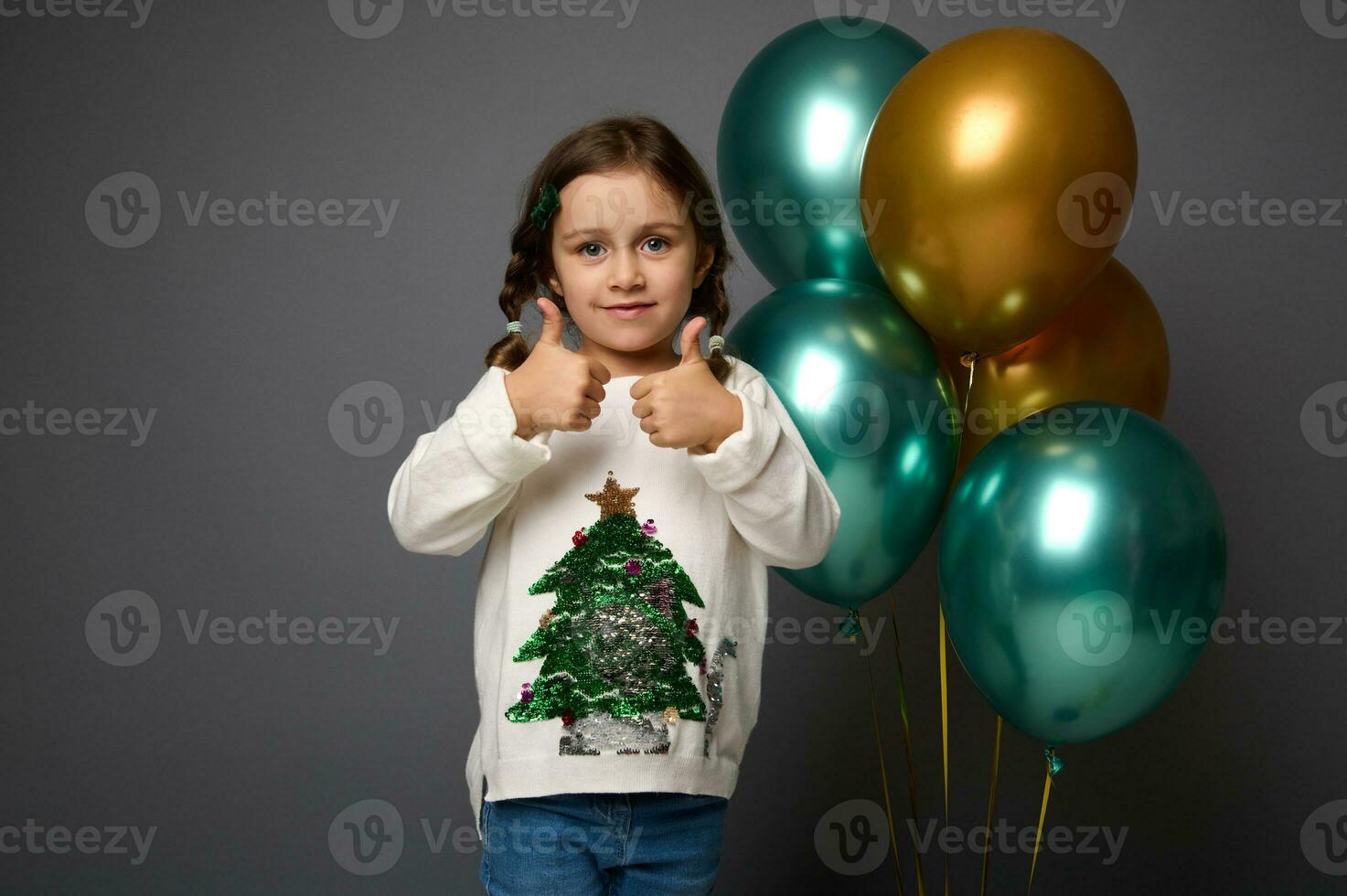 affascinante adorabile carino sorridente 4 anni vecchio bambino ragazza indossare bianca maglione mostrando pollici su, guardare a telecamera, isolato al di sopra di grigio sfondo con copia spazio per Natale e nuovo anno pubblicità foto