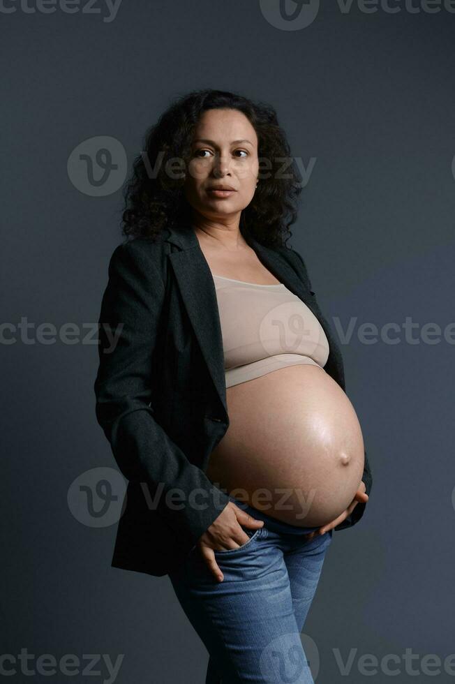 attraente incinta donna nel grigio blazer e beige reggiseno, Tenere gonfiarsi, guardare a parte, isolato su grigio studio sfondo foto