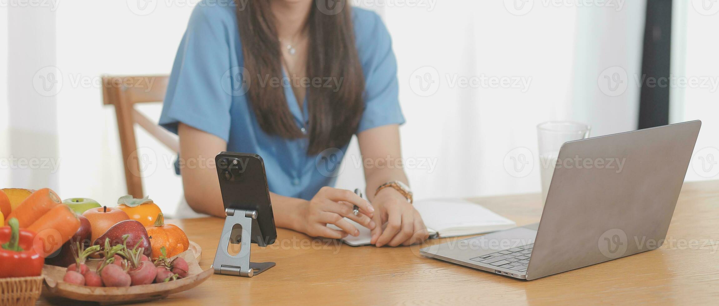 un' giovane donna con un' bellissimo viso nel un' blu camicia con lungo capelli mangiare frutta seduta dentro il cucina a casa con un' il computer portatile e taccuino per rilassamento, concetto vacanza. foto