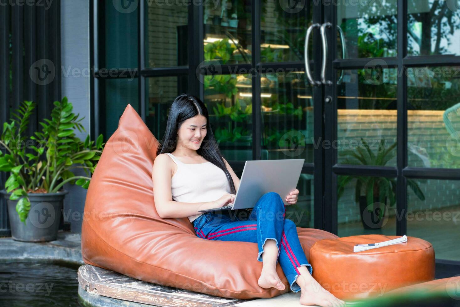 giovane adulto asiatico donna d'affari seduta su cuscino, Tenere penna e breve nota, guardare giù per sua il computer portatile nel davanti di porta di vetro di il bar durante sua lungo vacanza, digitale nomade di donna concetto. foto