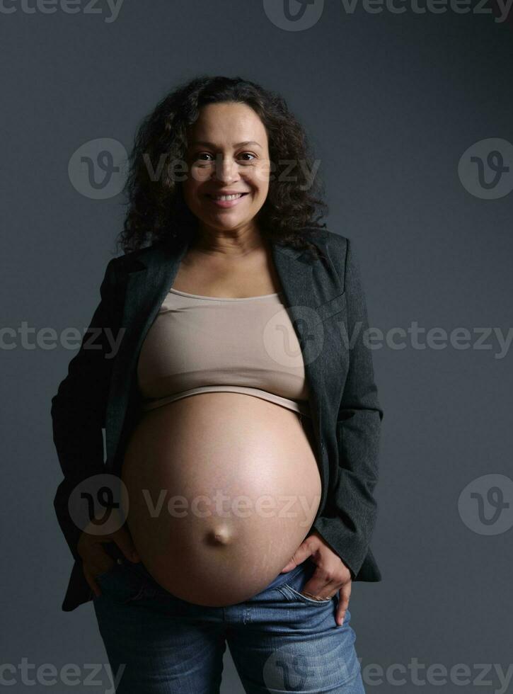 bellissimo africano americano donna, in attesa madre sorridente a telecamera, in posa spoglio incinta pancia al di sopra di grigio sfondo foto