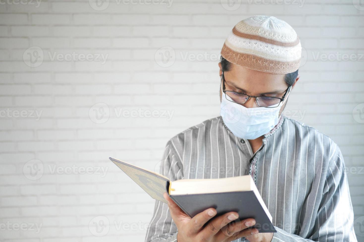 uomo musulmano con maschera facciale che legge il libro di agrifoglio del Corano al coperto foto
