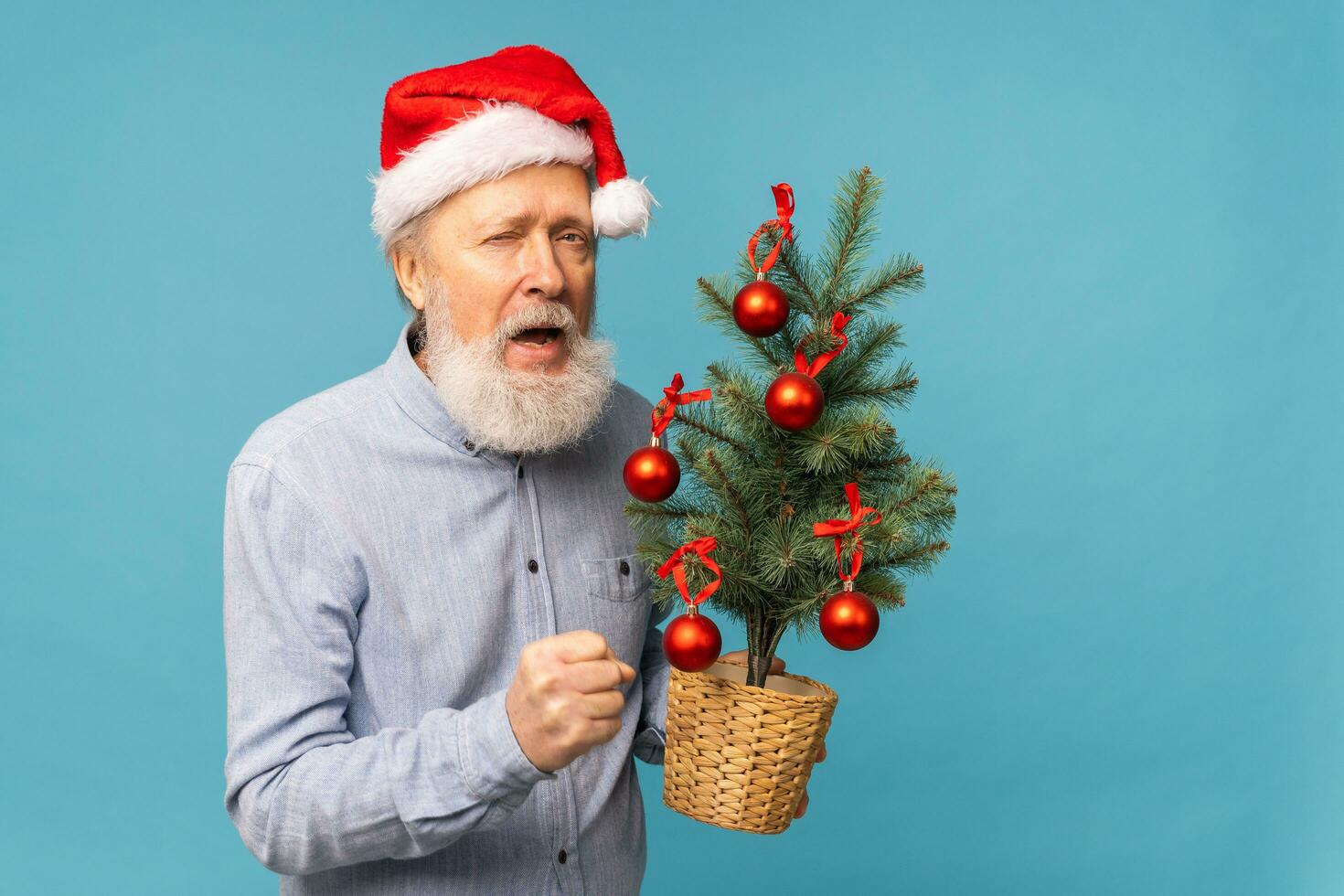 ritratto di contento emozioni Santa Claus eccitato guardare a telecamera e detiene piccolo Natale albero su blu sfondo con copia spazio foto