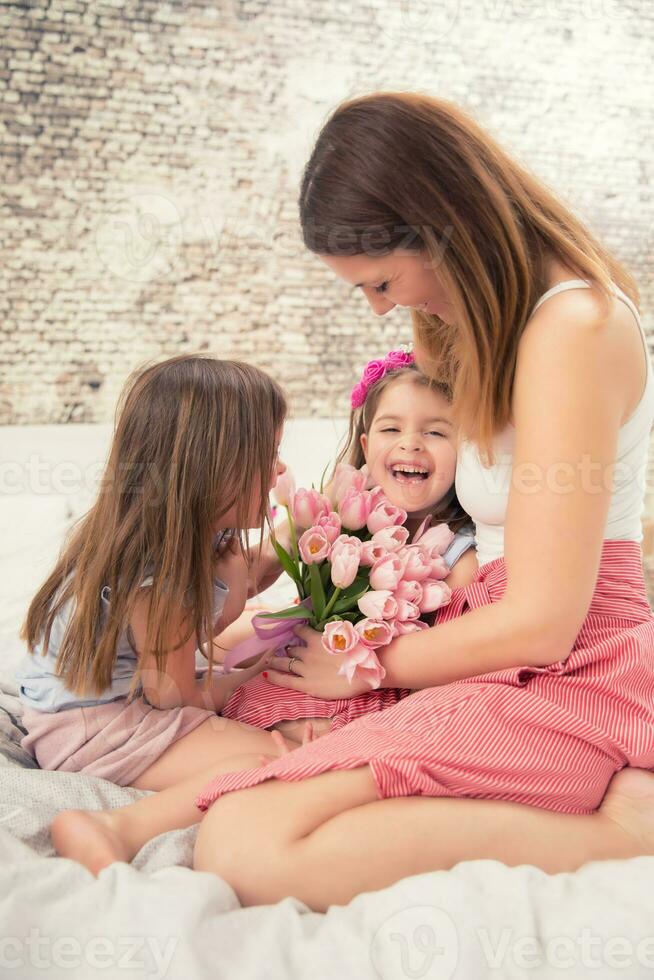 contento La madre di giorno concetto. mamma con twocute giovane figlie Gemelli su il letto nel il Camera da letto e un' mazzo di fiori tulipani foto