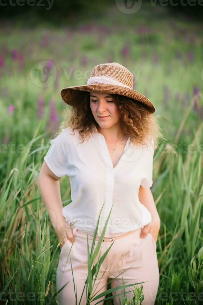 giovane donna sta nel bianca camicia nel campo di viola e rosa lupini. bellissimo giovane donna con Riccio capelli e cappello all'aperto su un' prato, lupini fiore. tramonto o Alba, luminosa sera leggero foto