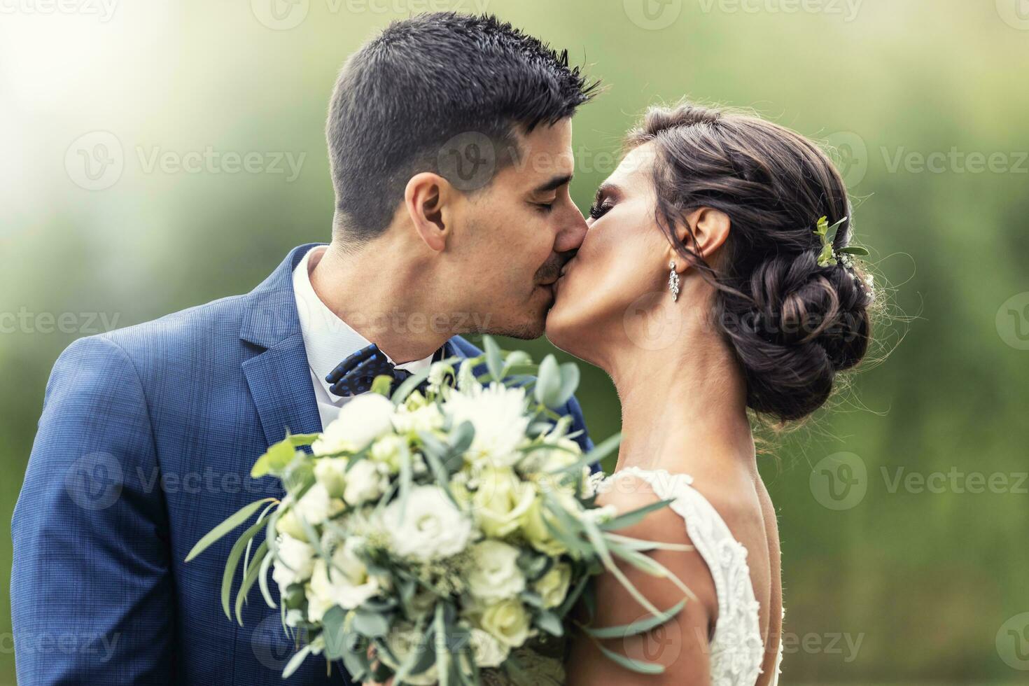 Novelli sposi bacio all'aperto mentre Tenere un' nozze mazzo di bianca fiori nel davanti di loro foto