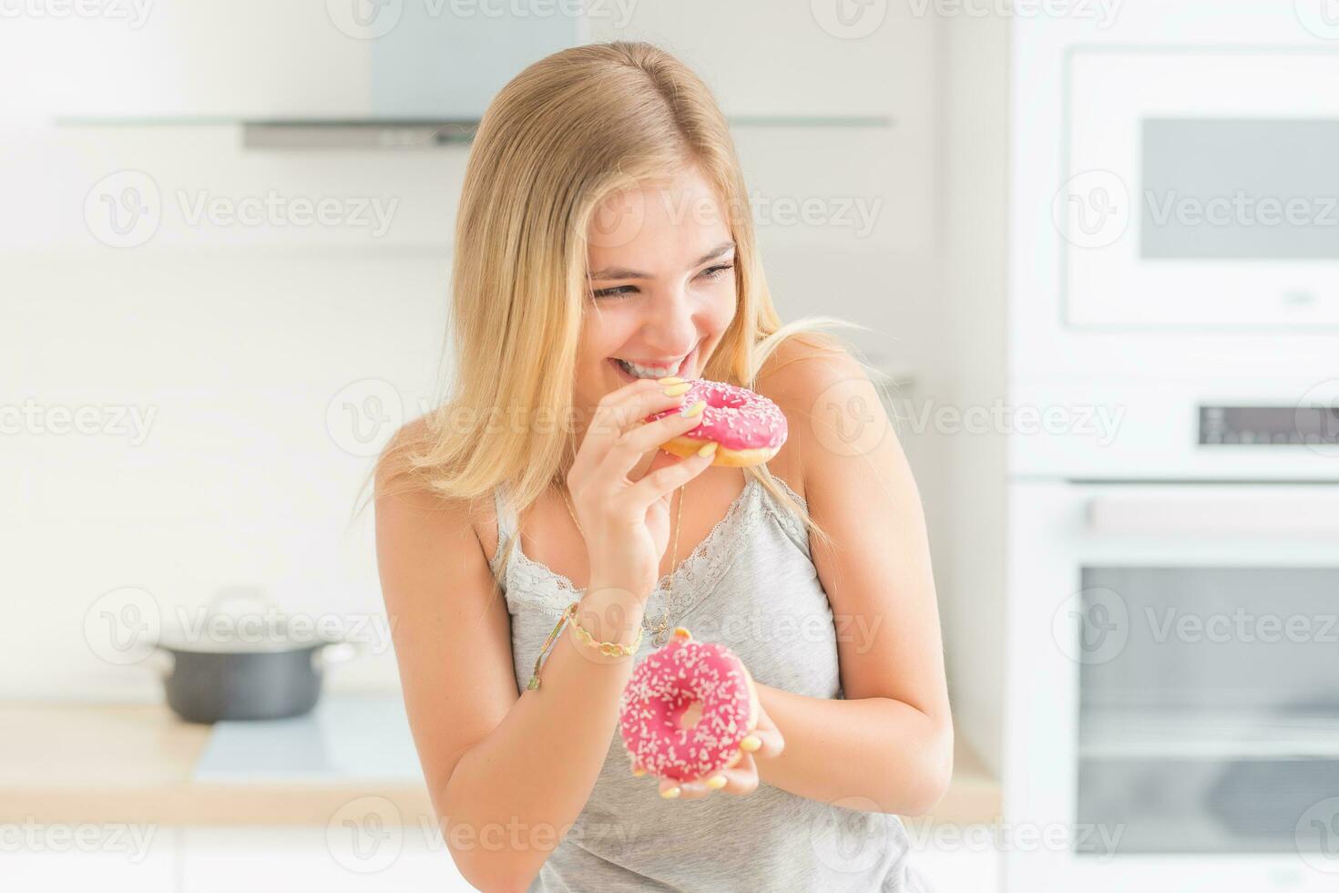 giovane bionda ragazza mangia rosa ciambelle nel casa cucina con gusto emozioni foto