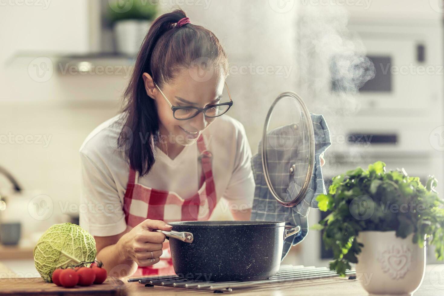 casalinga nel grembiule si apre coperchio di un' pentola guardare in esso mentre cucinando nel il cucina a casa foto